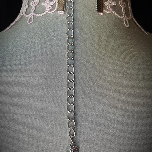 Puede incluir: Un collar gargantilla de encaje rosa con una cadena plateada y un colgante en forma de coraz&oacute;n. La gargantilla tiene un delicado dise&ntilde;o de encaje floral y un cierre met&aacute;lico. La cadena se extiende hacia abajo con un peque&ntilde;o dije de coraz&oacute;n.
