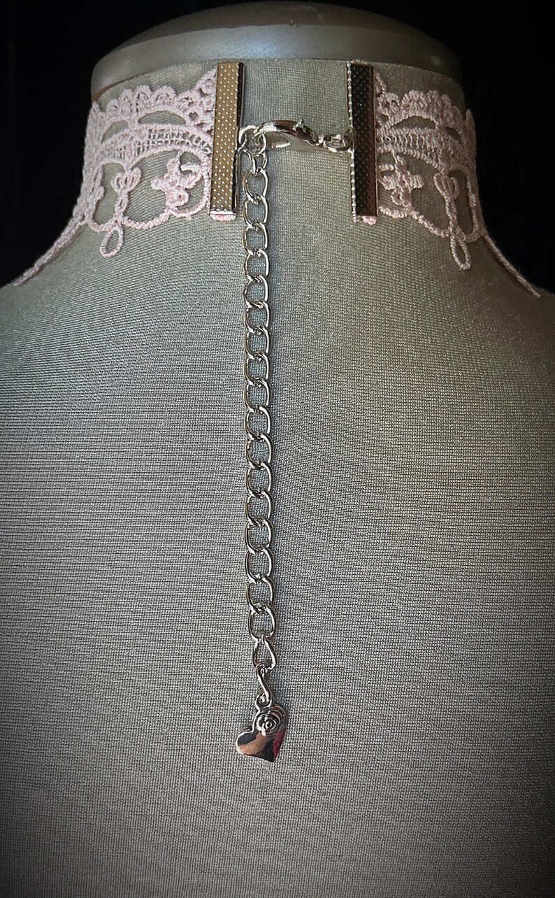 Puede incluir: Un collar gargantilla de encaje rosa con una cadena plateada y un colgante en forma de coraz&oacute;n. La gargantilla tiene un delicado dise&ntilde;o de encaje floral y un cierre met&aacute;lico. La cadena se extiende hacia abajo con un peque&ntilde;o dije de coraz&oacute;n.
