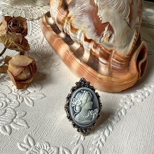 May include: A silver ring with a cameo of a woman in a white dress. The cameo is set in an oval frame with ornate details. The ring is on a white surface with a floral pattern.