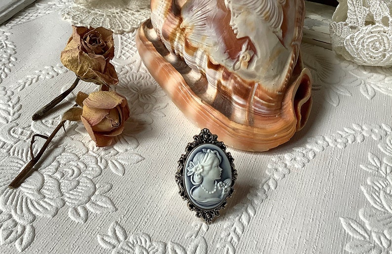 May include: A silver ring with a cameo of a woman in a white dress. The cameo is set in an oval frame with ornate details. The ring is on a white surface with a floral pattern.