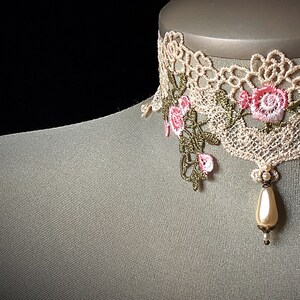 Puede incluir: Un delicado collar de encaje marfil con bordados florales rosas y verdes y un colgante de perla.