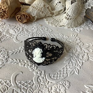 May include: Black metal bracelet with a white cameo featuring a woman's profile. The bracelet has a delicate, lacy design.