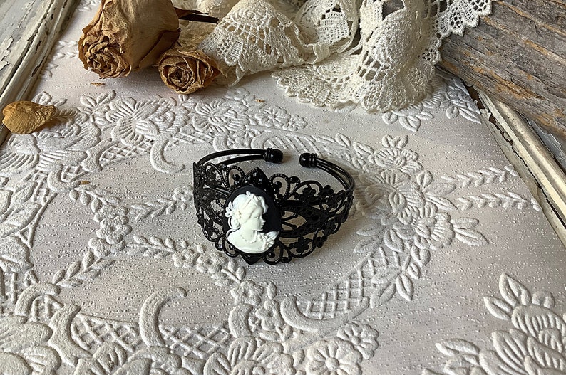 May include: Black metal bracelet with a white cameo featuring a woman's profile. The bracelet has a delicate, lacy design.