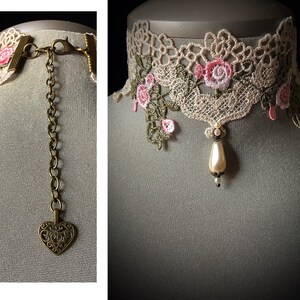 Puede incluir: Un delicado collar de encaje marfil con bordados florales rosas y un colgante de perla. El collar tiene una cadena de bronce con un dije en forma de coraz&oacute;n.