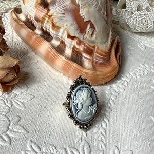 May include: A silver ring with a white cameo featuring a woman's profile. The cameo is set in an oval frame with intricate details. The ring is on a white surface with a floral pattern.