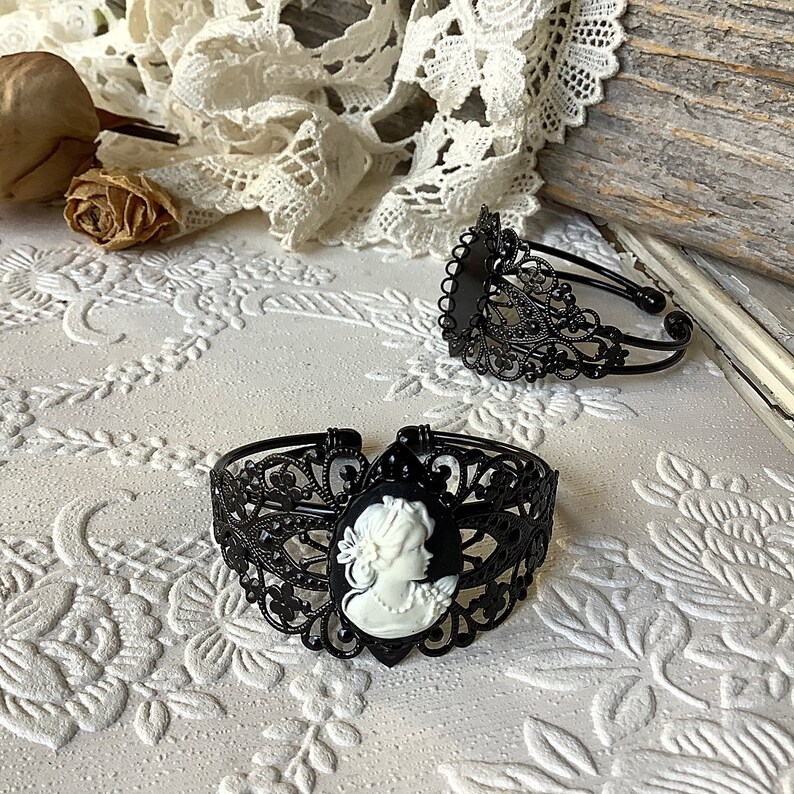 May include: Two black metal bracelets with intricate filigree designs. Each bracelet features a cameo of a woman's profile in white.
