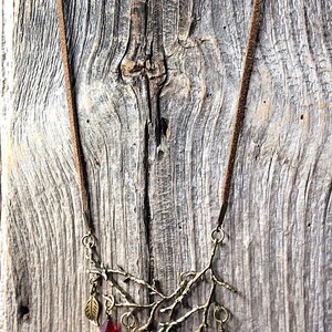 May include: A bronze-toned necklace featuring a branch design with a brown suede cord. The branch is adorned with colorful, faceted crystal beads in red, orange, yellow, green, blue, and purple. A small leaf charm hangs from the branch.