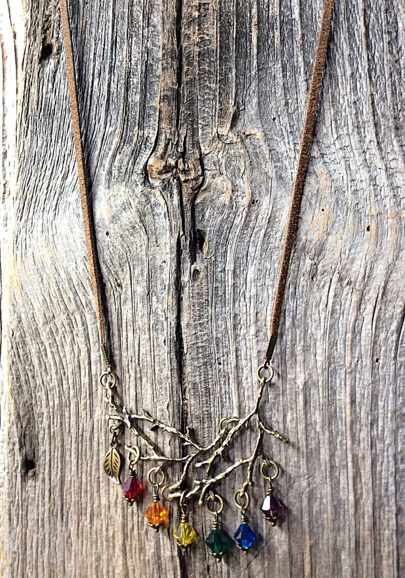 May include: A bronze-toned necklace featuring a branch design with a brown suede cord. The branch is adorned with colorful, faceted crystal beads in red, orange, yellow, green, blue, and purple. A small leaf charm hangs from the branch.