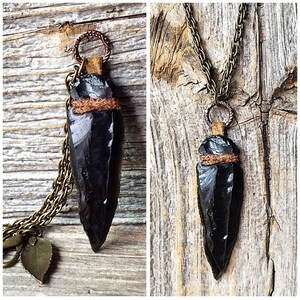 Puede incluir: Un collar con un colgante de punta de flecha de obsidiana negra, con una envoltura de cuero marr&oacute;n y una cadena de lat&oacute;n.