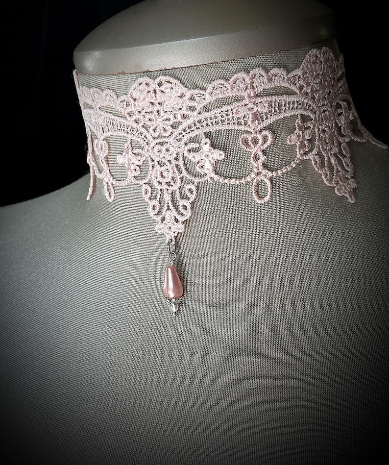 Puede incluir: Un delicado collar de gargantilla de encaje rosa claro. El intrincado dise&ntilde;o de encaje presenta patrones florales y geom&eacute;tricos. Un colgante de perla rosa en forma de l&aacute;grima cuelga del centro. El collar se exhibe en un maniqu&iacute;.