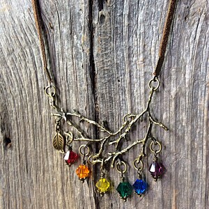 May include: A bronze-toned necklace featuring a branch design with colorful crystal pendants. The necklace has a brown suede cord and a leaf charm. The crystals are red, orange, yellow, green, blue, and purple.