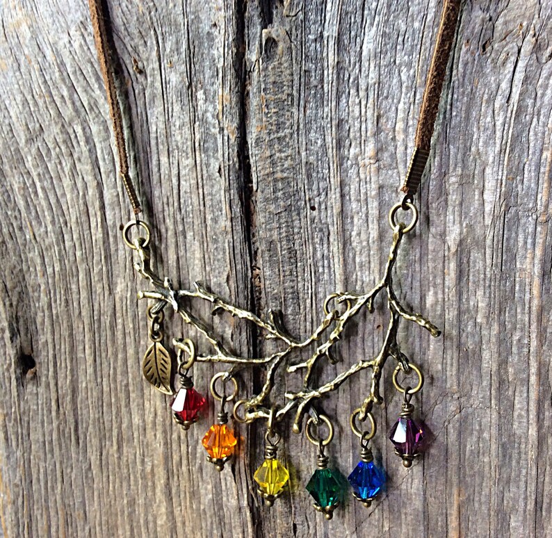 May include: A bronze-toned necklace featuring a branch design with colorful crystal pendants. The necklace has a brown suede cord and a leaf charm. The crystals are red, orange, yellow, green, blue, and purple.