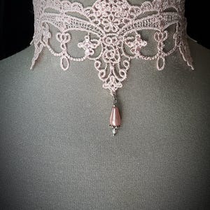 Puede incluir: Un delicado collar gargantilla de encaje rosa claro. El intrincado dise&ntilde;o de encaje presenta patrones florales y de volutas. Una cuenta rosa en forma de l&aacute;grima cuelga del centro. El collar se exhibe en un maniqu&iacute;.