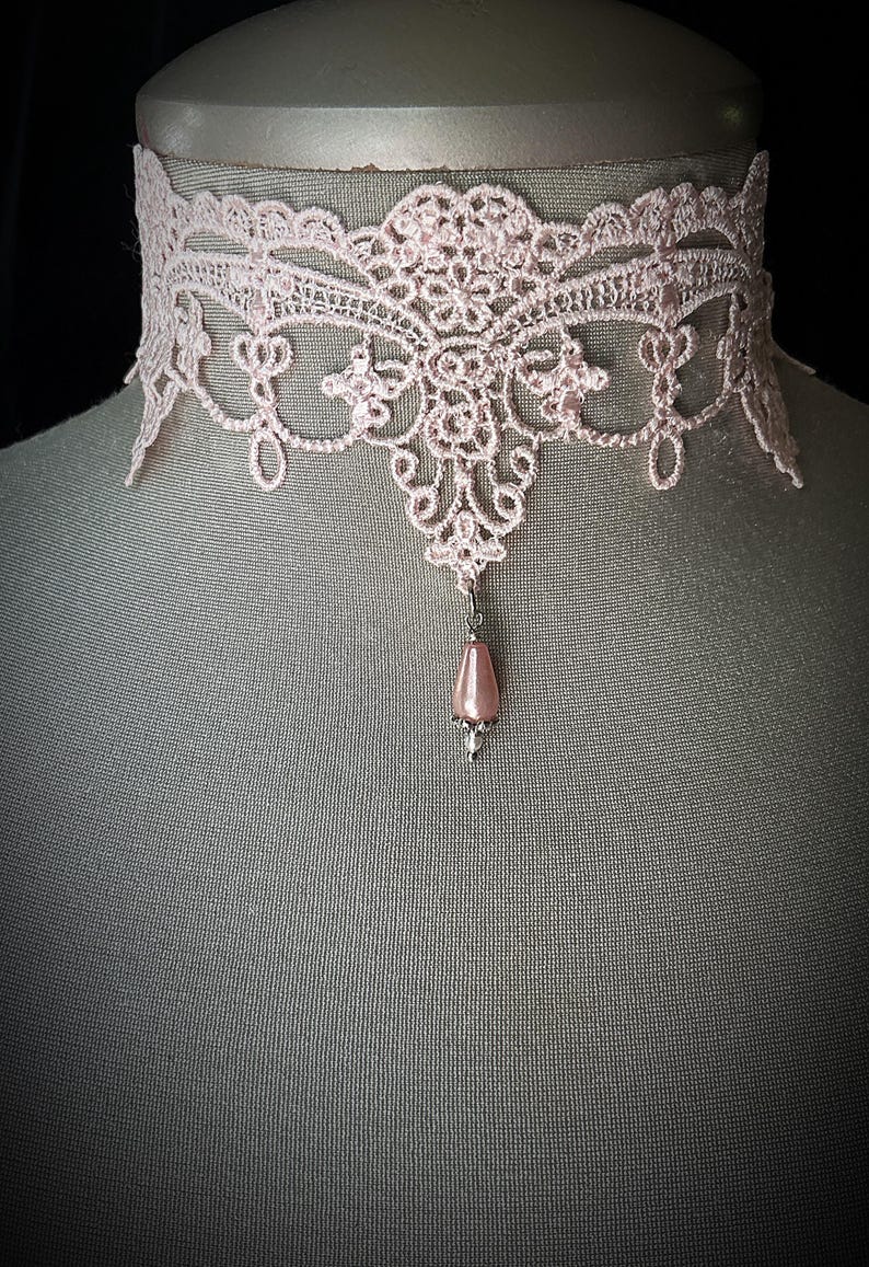 Puede incluir: Un delicado collar gargantilla de encaje rosa claro. El intrincado dise&ntilde;o de encaje presenta patrones florales y de volutas. Una cuenta rosa en forma de l&aacute;grima cuelga del centro. El collar se exhibe en un maniqu&iacute;.