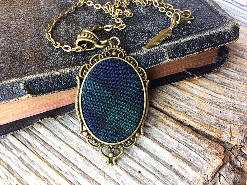 May include: A gold-toned necklace with a dark blue and green plaid fabric pendant in an oval frame.