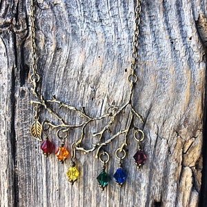 May include: A bronze-toned necklace featuring a branch design with a leaf pendant. The branch has six colorful, faceted crystal pendants in red, orange, yellow, green, blue, and purple. The necklace is displayed against a weathered wood background.