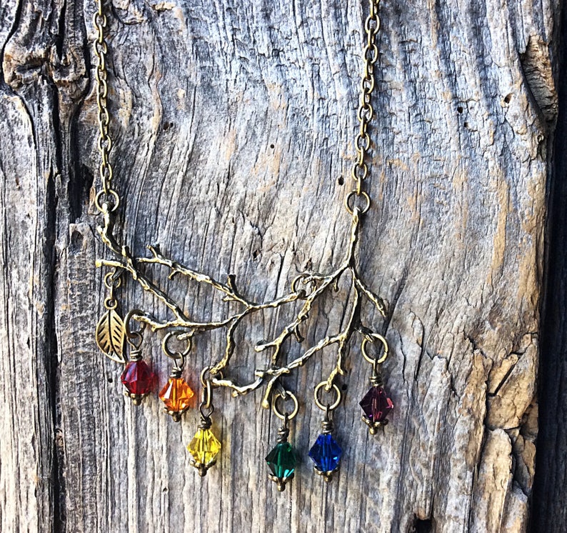 May include: A bronze-toned necklace featuring a branch design with a leaf pendant. The branch has six colorful, faceted crystal pendants in red, orange, yellow, green, blue, and purple. The necklace is displayed against a weathered wood background.