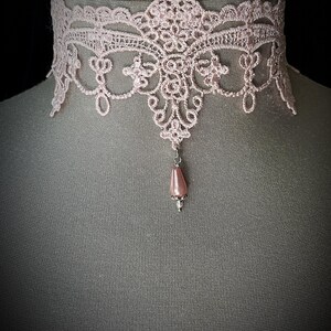 Puede incluir: Un delicado collar gargantilla de encaje rosa claro. El intrincado dise&ntilde;o de encaje presenta patrones florales y de volutas. Un colgante en forma de l&aacute;grima, con una perla rosa, cuelga del centro. El collar se exhibe en un maniqu&iacute;.
