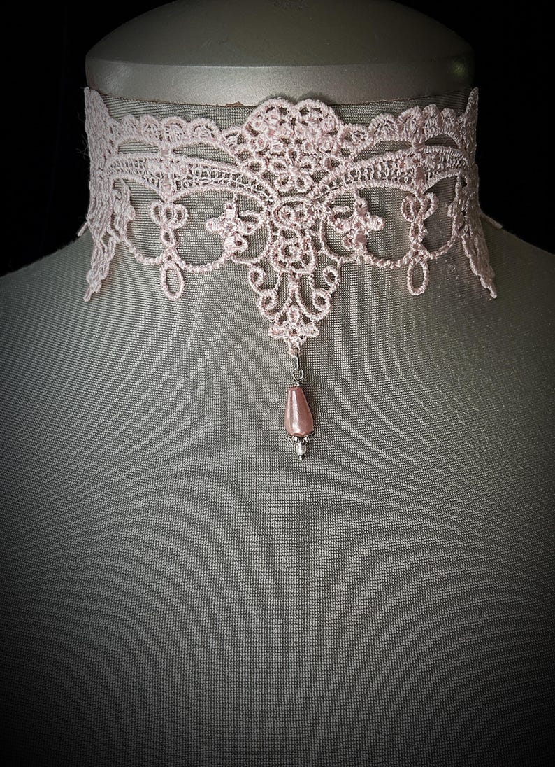 Puede incluir: Un delicado collar gargantilla de encaje rosa claro. El intrincado dise&ntilde;o de encaje presenta patrones florales y de volutas. Un colgante en forma de l&aacute;grima, con una perla rosa, cuelga del centro. El collar se exhibe en un maniqu&iacute;.