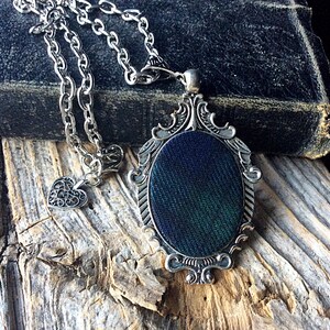 May include: A silver chain necklace with a heart charm and a pendant featuring a dark green and blue fabric design in an ornate silver frame.