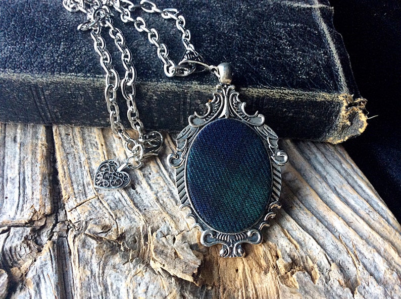 May include: A silver chain necklace with a heart charm and a pendant featuring a dark green and blue fabric design in an ornate silver frame.
