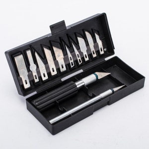 May include: A black plastic case with a set of 12 silver utility knives and two additional silver utility knives. The case has a black handle and a silver blade.