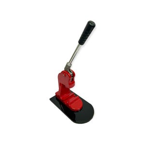 May include: A red button maker with a black base and a silver lever. The handle is black and textured. This machine is designed for creating buttons and badges.