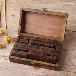 May include: A wooden box with a hinged lid, filled with individual wooden letter stamps. The stamps include uppercase and lowercase letters, numbers, and punctuation marks. The box and stamps are a warm brown color.