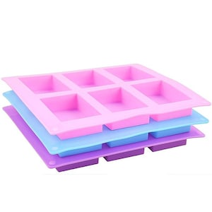 May include: Three colourful silicone soap moulds stacked on top of each other. The top mould is pink, the middle one is light blue, and the bottom one is purple. Each mould has six rectangular cavities.