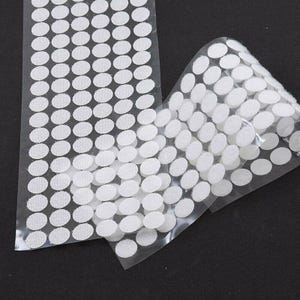 May include: Two sheets of white, oval-shaped hook and loop fasteners on a clear backing. The fasteners are arranged in neat rows and columns. The background is a solid black colour.