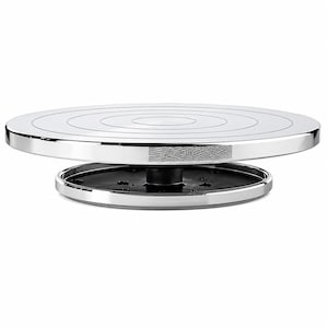 May include: A circular rotating display stand featuring a polished chrome finish on the top surface with concentric etched rings. The base is constructed from black plastic with a chrome-finished outer rim and a textured grip section on the side.