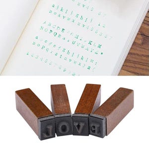 May include: A set of four wooden stamps spelling "LOVE" with individual letter stamps. The stamps have dark brown wooden handles and black rubber letter faces. The image also shows a page with printed green letters and numbers.