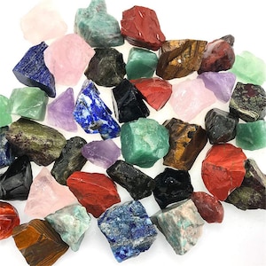 May include: A selection of raw gemstones in a variety of colours, including pink, green, red, blue, and black. The stones have a rough, natural texture and are scattered against a white background. The assortment includes amethyst, lapis lazuli, and rose quartz.