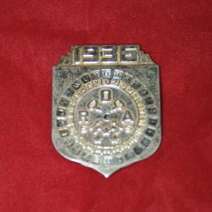 Puede incluir: Una insignia de metal plateada con el año 1935 y las letras "R.O.A." en el centro. La insignia tiene forma de escudo y un borde decorativo.