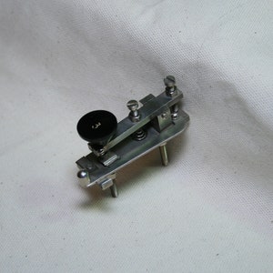 May include: A silver metal telegraph key with a black plastic knob. The key has two metal posts and a spring.