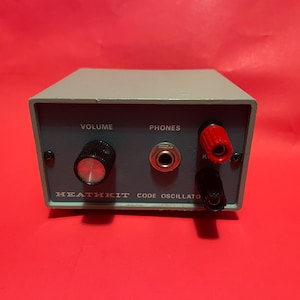 May include: A vintage Heathkit Code Oscillator, a grey rectangular device with a dark grey faceplate. It features a volume knob, a headphone jack, and two terminals. The text "VOLUME", "PHONES", and "HEATHKIT CODE OSCILLATOR" are visible.