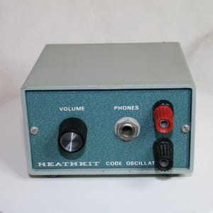 Puede incluir: Un oscilador de código Heathkit gris con un panel frontal color verde azulado. El panel cuenta con una perilla de volumen negra, un conector para auriculares plateado y bornes rojo y negro. El texto dice VOLUME, PHONES y HEATHKIT CODE OSCILLATOR.