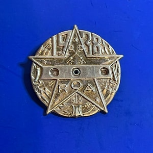 Può includere: Un distintivo in metallo argentato con una stella a cinque punte al centro. La stella contiene le lettere "ROA" all'interno e l'anno "1928" intorno all'esterno. Il distintivo ha un design a corona intorno all'esterno.