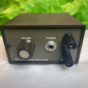 Puede incluir: Un dispositivo electrónico gris oscuro con una perilla de volumen, un conector para auriculares y dos perillas más pequeñas. El texto "VOLUME", "PHONES" y "Hear... CODE OSCILLATOR" es visible en el panel frontal. El dispositivo tiene forma rectangular.