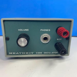 Puede incluir: Un oscilador de código Heathkit con una carcasa rectangular gris y un panel frontal verde. Cuenta con una perilla de volumen negra, un conector central para auriculares y dos bornes de conexión etiquetados como KEY, uno rojo y otro negro.