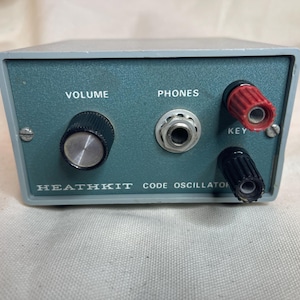 May include: A gray Heathkit Code Oscillator electronic device with a teal front panel. It features a black volume knob, a central headphone jack labeled PHONES, and two binding posts labeled KEY, one red and one black, for telegraphy practice.
