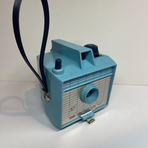 May include: A light blue vintage Savoy box camera featuring a grid-patterned front plate with the text SAVOY and a small planet graphic. The camera includes a dark blue top-mounted knob, a central lens, and a black plastic wrist strap attached to the side.