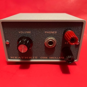 May include: A grey Heathkit Code Oscillator with a dark grey front panel. It has a volume knob, a phones jack, and key terminals. The text "VOLUME", "PHONES", and "KEY" are visible.