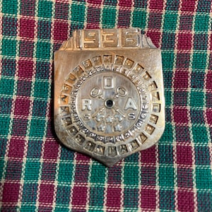 Puede incluir: Un calendario de metal vintage con el año 1936 en la parte superior. El calendario tiene un dial circular con números y letras, y un diseño central con estrellas y símbolos. El metal tiene una apariencia desgastada y envejecida.