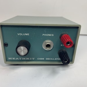 Puede incluir: Un oscilador de código Heathkit gris con un panel frontal azul verdoso. El panel cuenta con una perilla de volumen negra, un conector para auriculares central y terminales de llave rojos y negros. El texto dice VOLUME, PHONES, KEY y HEATHKIT CODE OSCILLATOR.