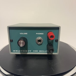 May include: A vintage Heathkit Code Oscillator, a grey and teal electronic device. It features a volume knob, a phones jack, and key terminals. The text "VOLUME", "PHONES", "KEY", and "HEATHKIT CODE OSCILLATO" are visible.