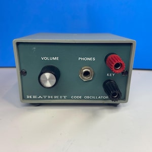 Puede incluir: Un oscilador de código Heathkit gris con un panel frontal que presenta una perilla de volumen negra, un conector central para auriculares etiquetado como PHONES y dos bornes de conexión rojo y negro etiquetados como KEY.