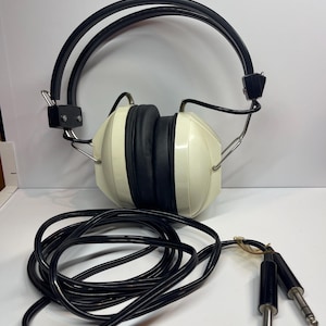May include: Vintage-style over-ear headphones featuring cream-coloured ear cups with black padding. The device includes a dual-wire black headband frame and a long black cord that terminates in two separate silver-tipped audio jack connectors.
