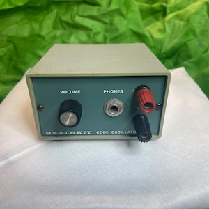 Puede incluir: Un oscilador de código Heathkit vintage en una carcasa metálica verde claro. El panel frontal es verde oscuro con una perilla de volumen, un conector para auriculares y dos bornes. El texto "Heathkit Code Oscillator" está impreso en el frente.
