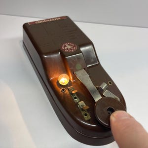 May include: A brown Brumberger Codemaster telegraph key device with a metallic lever and a glowing orange light. The top surface features printed text and a circular logo. The device is constructed from a dark brown moulded material with silver-toned metal components and a circular knob at the end of the lever.